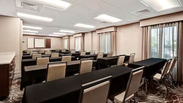 Une salle de conférence équipée de plusieurs tables et chaises, d’un pupitre et de grandes fenêtres avec des rideaux.