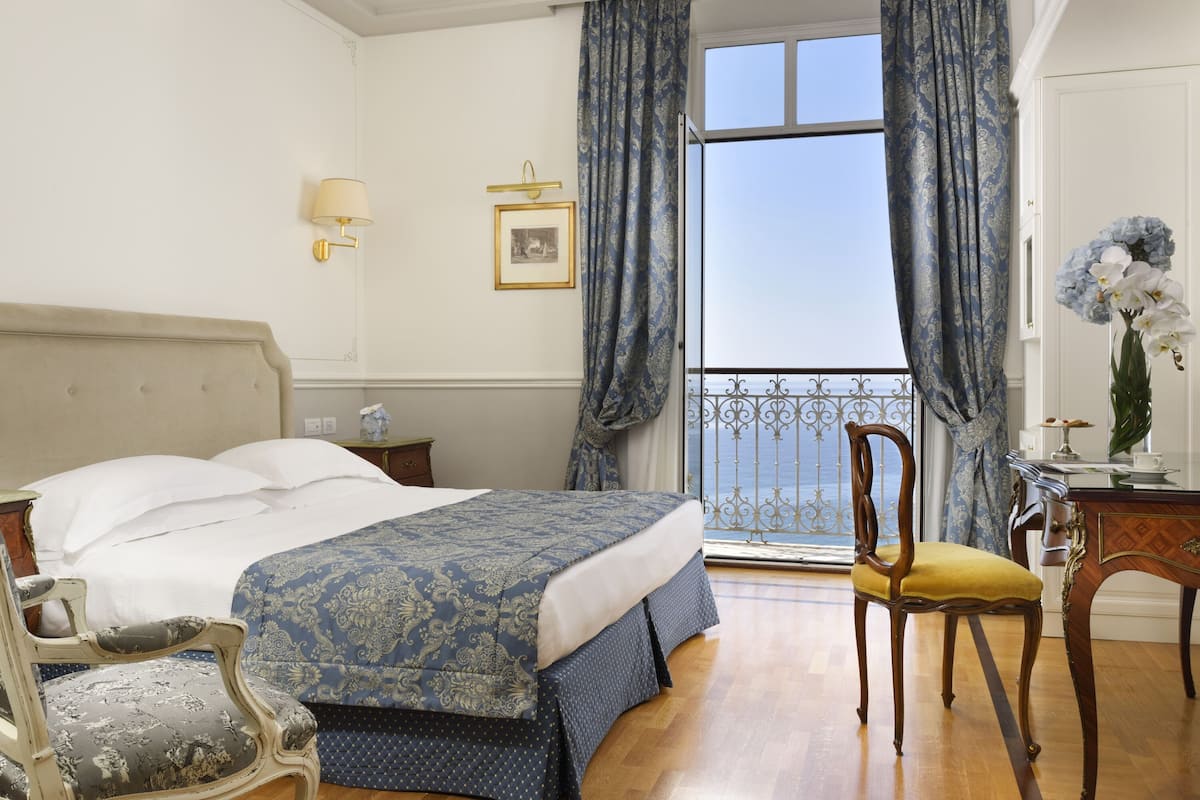 A hotel room with a bed, a chair, a desk, and a view of the sea through large windows.