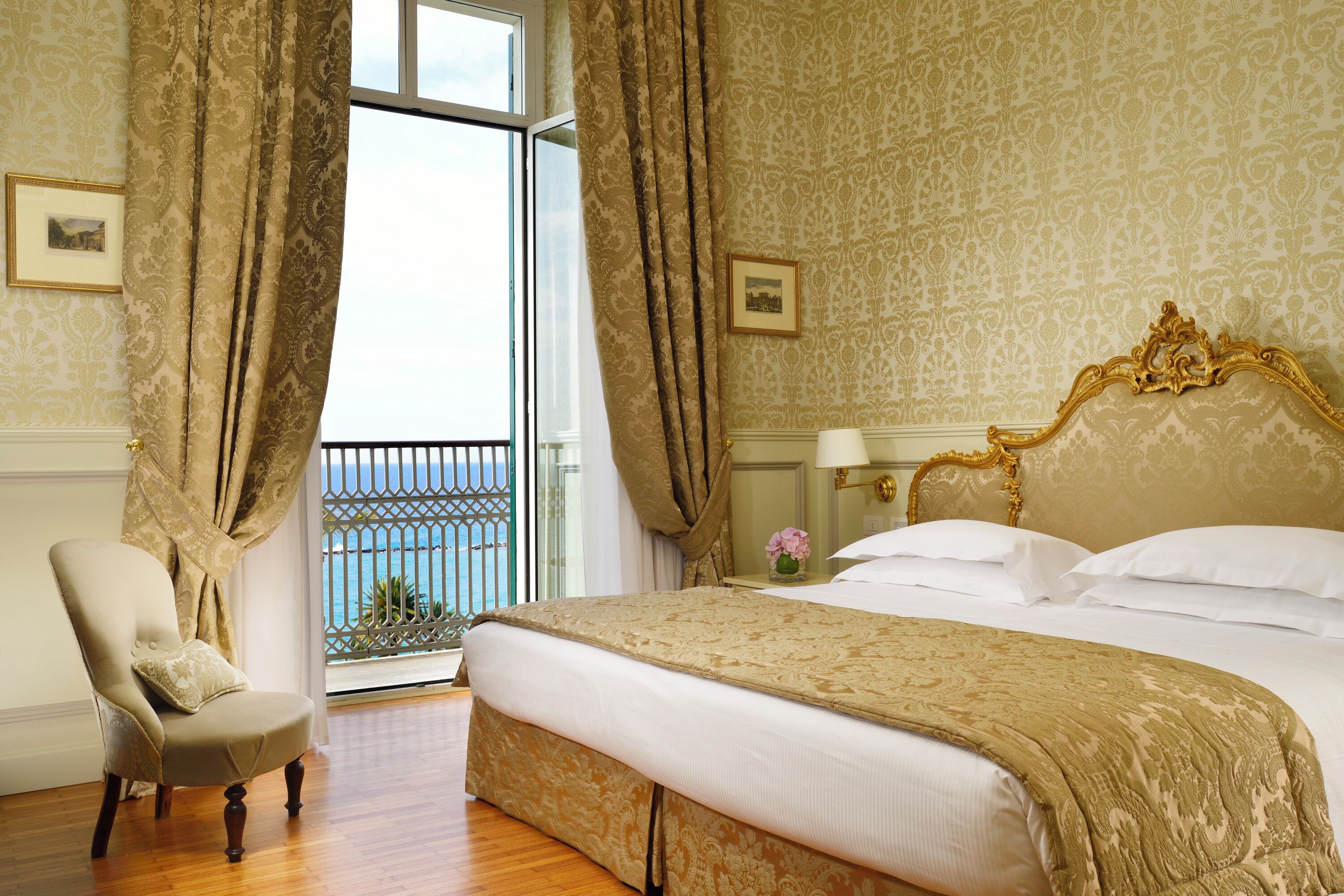 Habitación de hotel con una cama grande, una silla, una mesita de noche y vistas al mar desde un balcón.