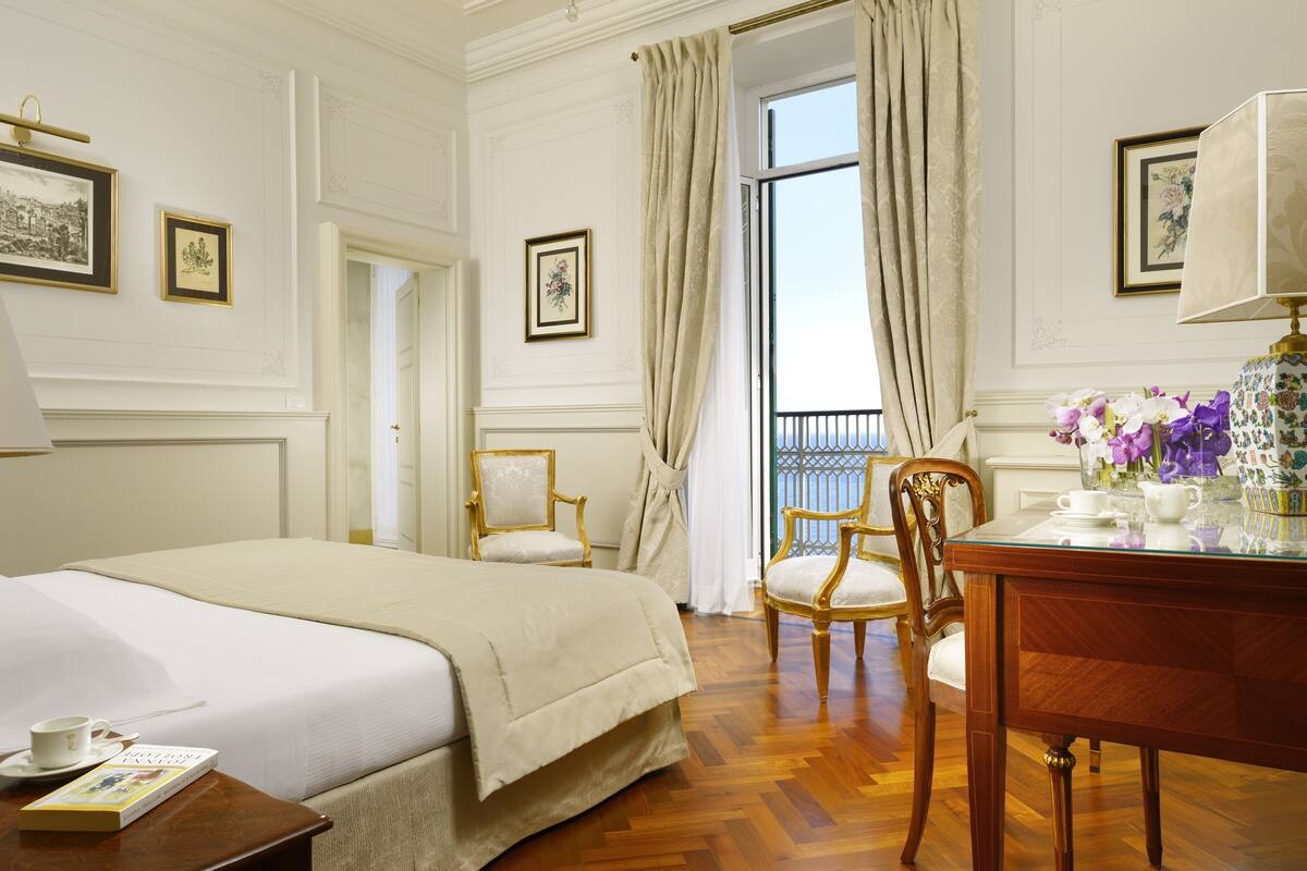 A hotel room with a large bed, two chairs, a table with a lamp, and framed pictures on the walls.