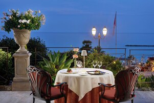 Un área de comedor al aire libre con una mesa para dos, rodeada de sillas, una planta en maceta y vista al mar.