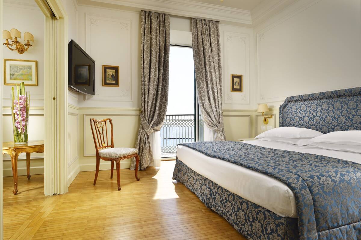 A hotel room with a large bed, a wooden chair, a TV mounted on the wall, and a balcony with curtains.