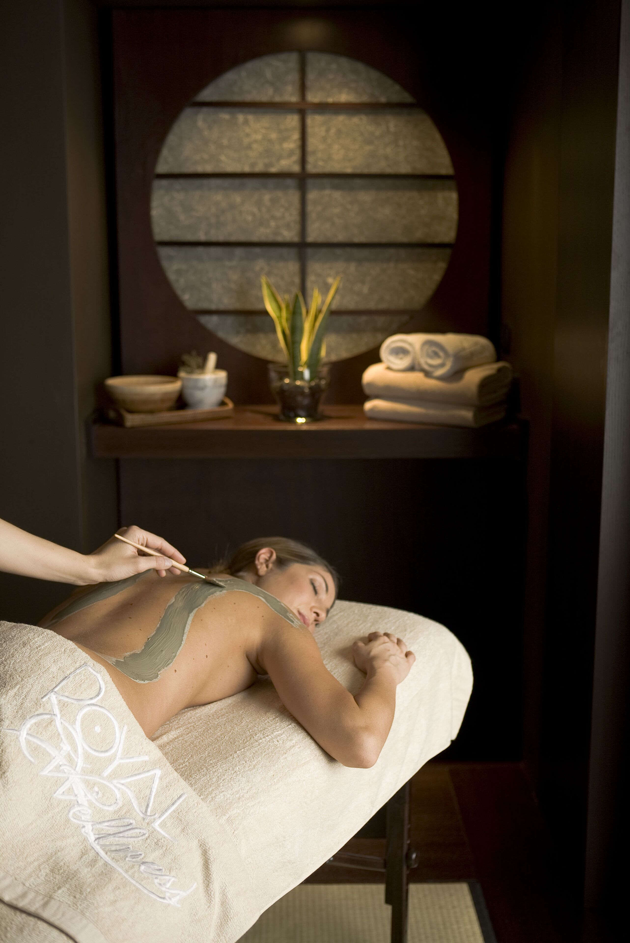 Ein Spa-Behandlungsraum mit Massagetisch, einem Handtuch mit dem Namen des Spas, einer Wand mit Rundfenster und einem Regal mit Handtüchern und einer Pflanze.