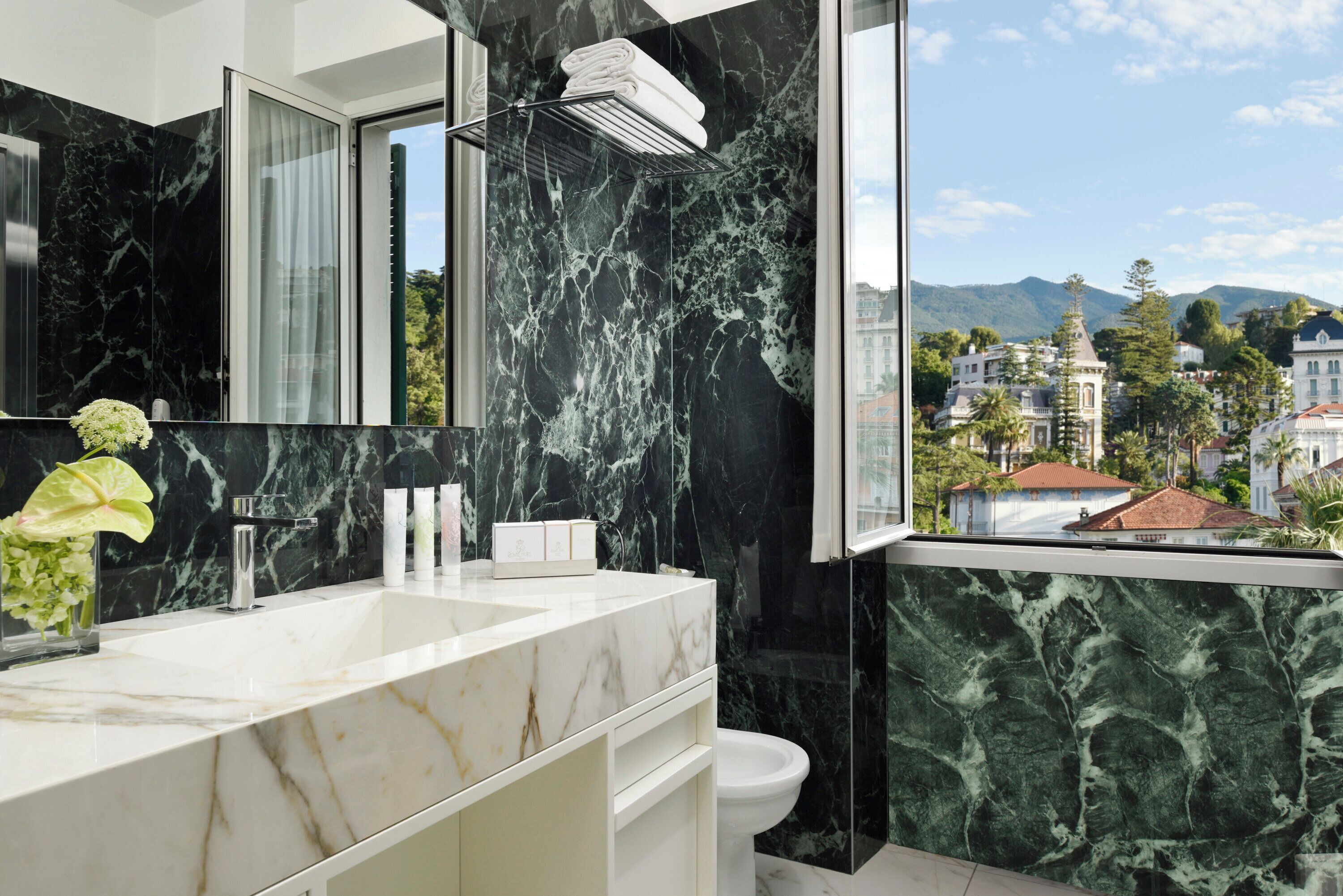 Une salle de bain moderne avec un grand lavabo en marbre, une douche en verre et une vue sur un paysage urbain avec des bâtiments et des montagnes.