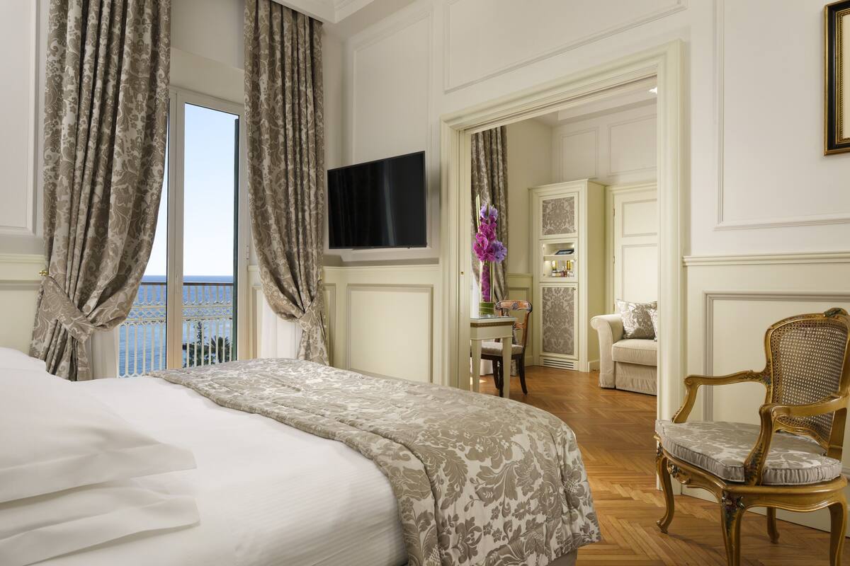 A hotel room with a large bed, a television, a desk with a chair, a sofa, and a view of the sea.