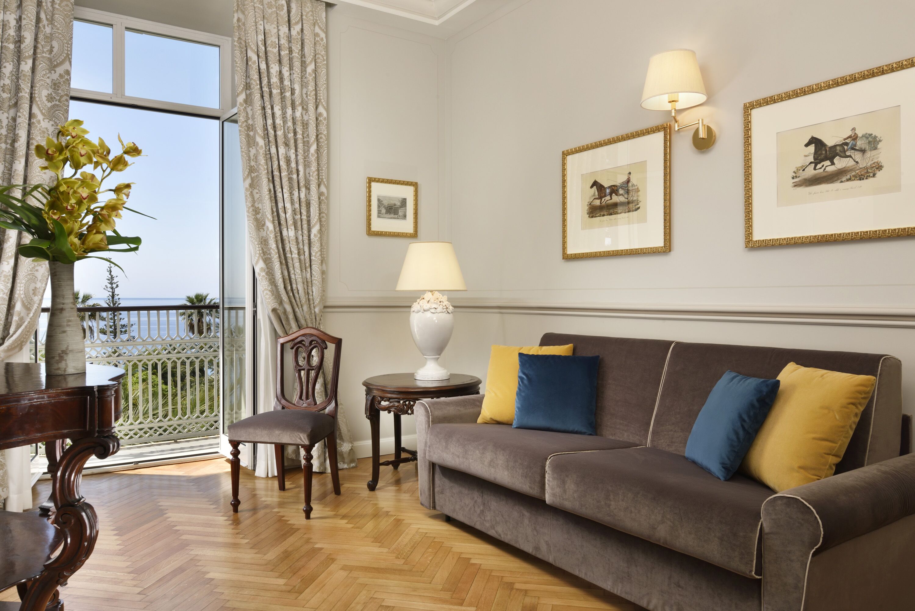 Un salon comprenant un canapé, des fauteuils, une table et une lampe. Des tableaux encadrés ornent le mur, et une fenêtre offre une vue sur l’extérieur.