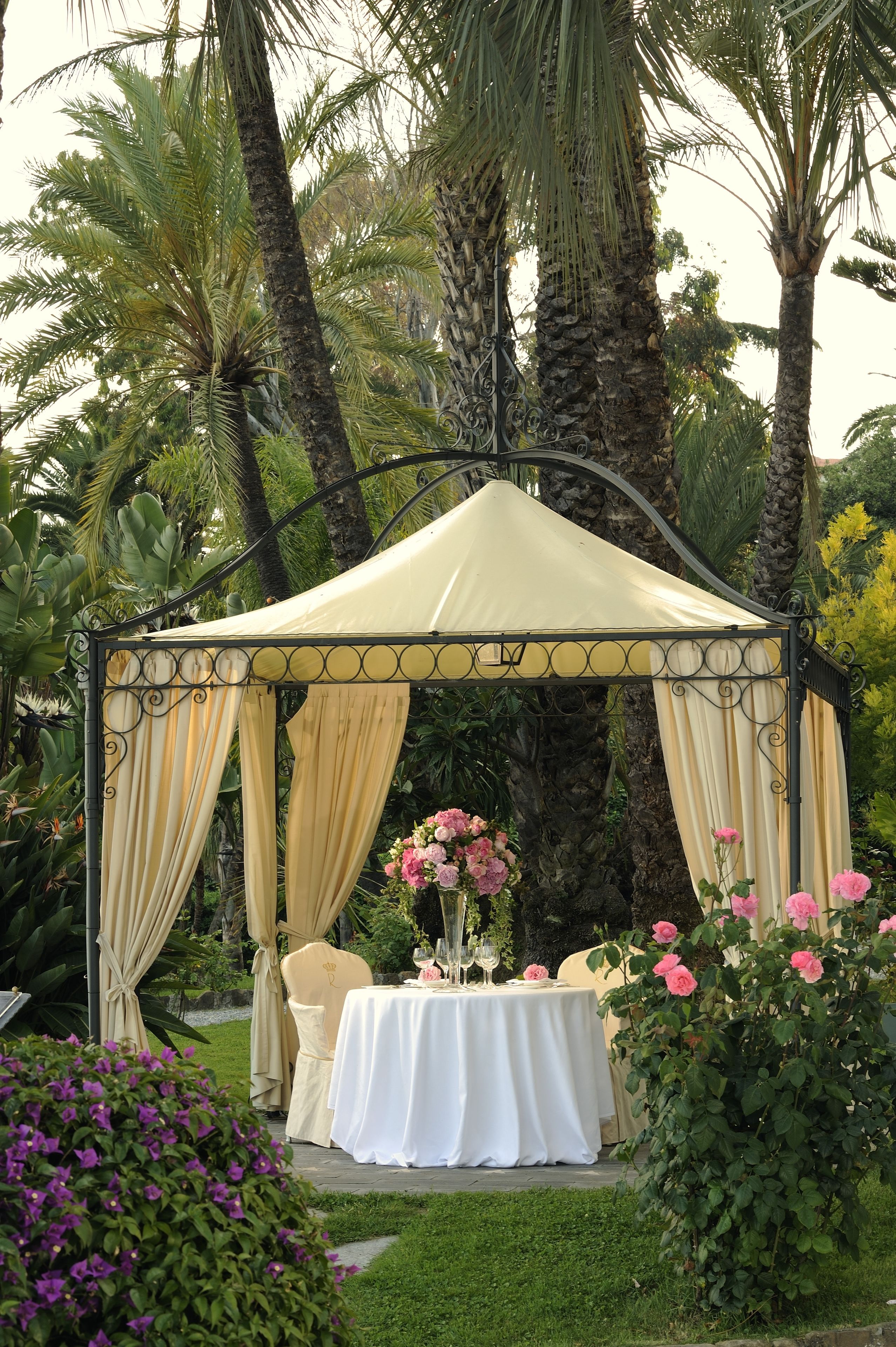 Un área de comedor al aire libre techada, con una mesa dispuesta para dos personas, rodeada de abundante follaje y palmeras.