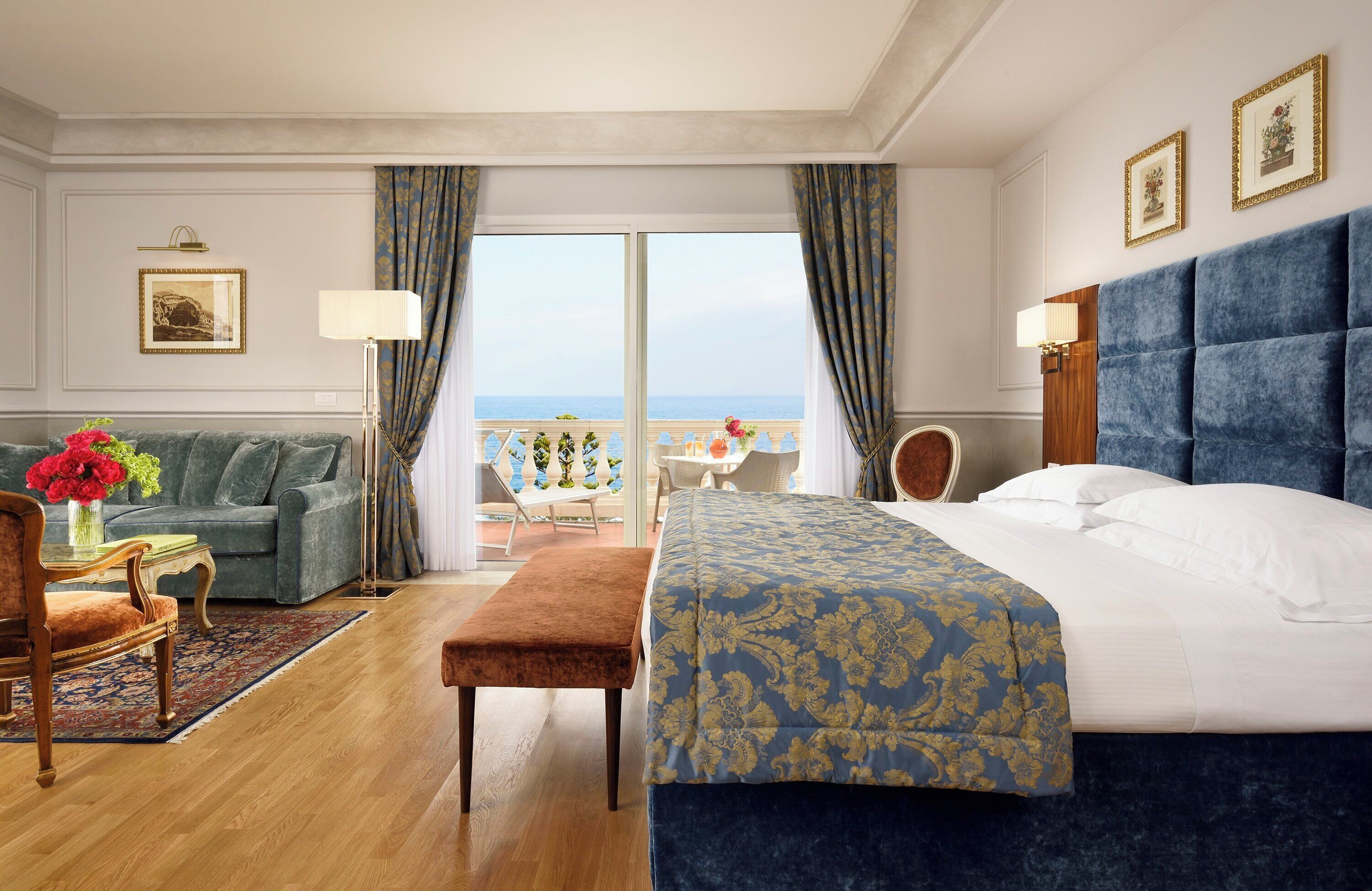 A hotel room with a large bed, a sofa, a small table with a vase of flowers, and a view of the sea through a large window.