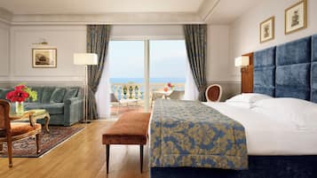 A hotel room with a large bed, a sofa, a small table with a vase of flowers, and a view of the sea through a large window.