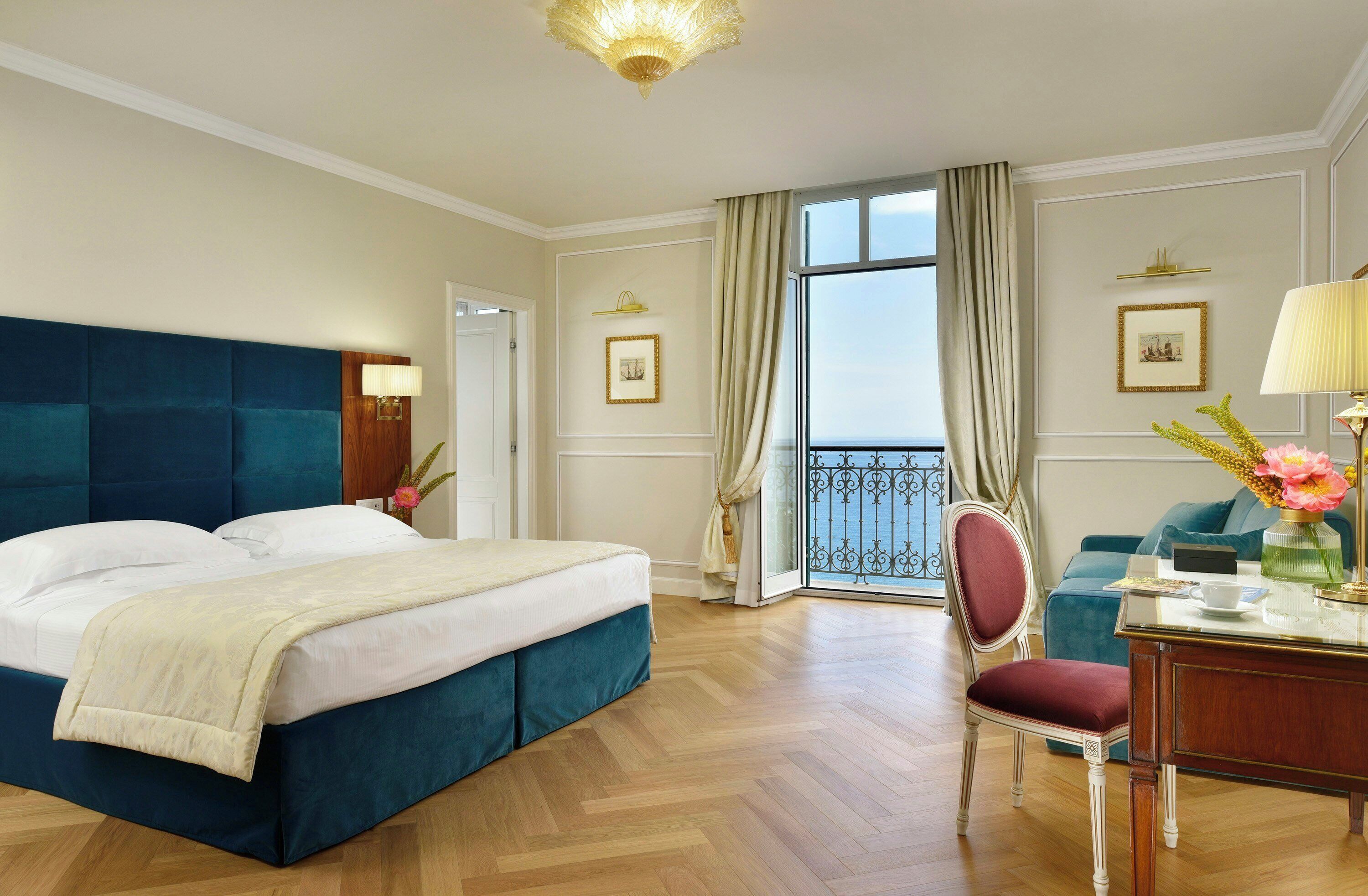 A hotel room with a large bed, a desk with a chair, a view of the sea through a balcony, and a chandelier.