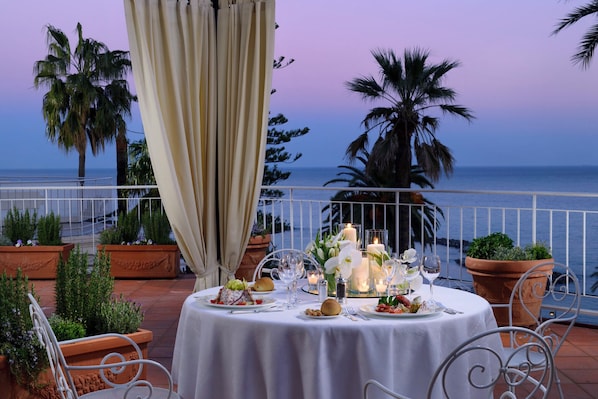 Ein Tisch, reichhaltig gedeckt, mit Blick auf das Meer, umgeben von Blumentöpfen und einem Geländer.