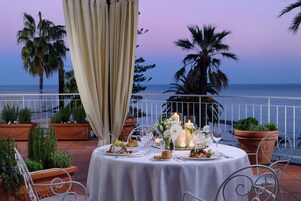 Una mesa dispuesta para comer, con vista al mar, rodeada de plantas en macetas y una barandilla.