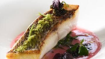 A dish with a piece of fish, green topping, and a red sauce on a white plate.