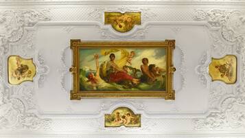 A ceiling with ornate white plasterwork and gold-framed paintings depicting classical scenes.