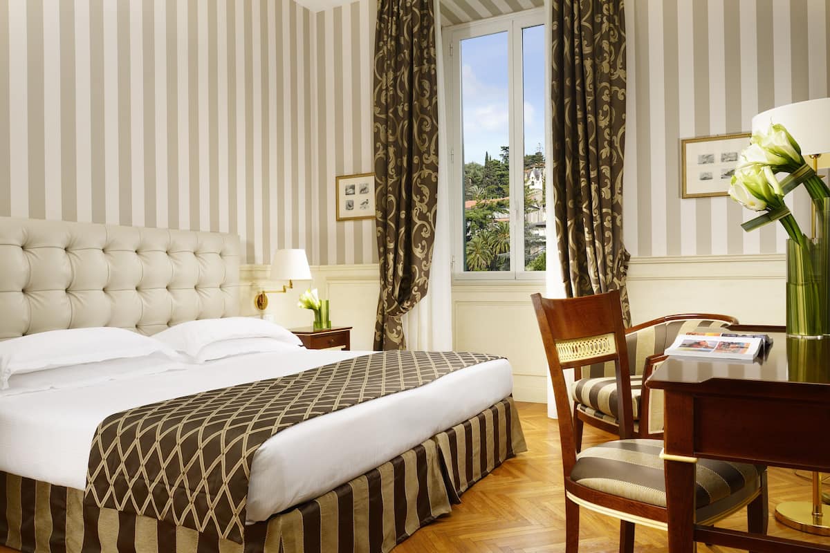 A hotel room with a large bed, a wooden chair, a table with a vase of flowers, and a window with curtains.