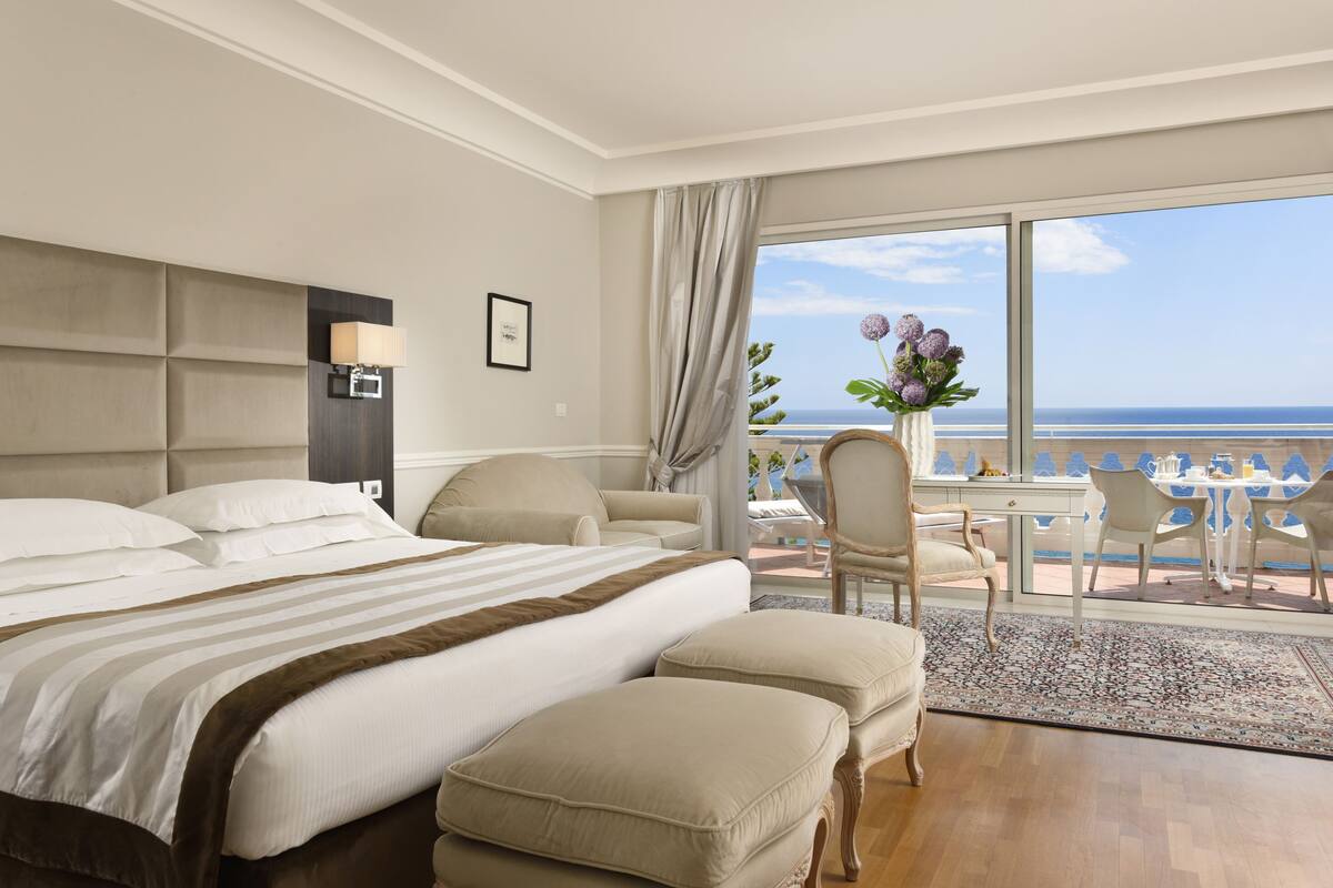 A hotel room with a large bed, a desk with chairs, and a view of the beach.