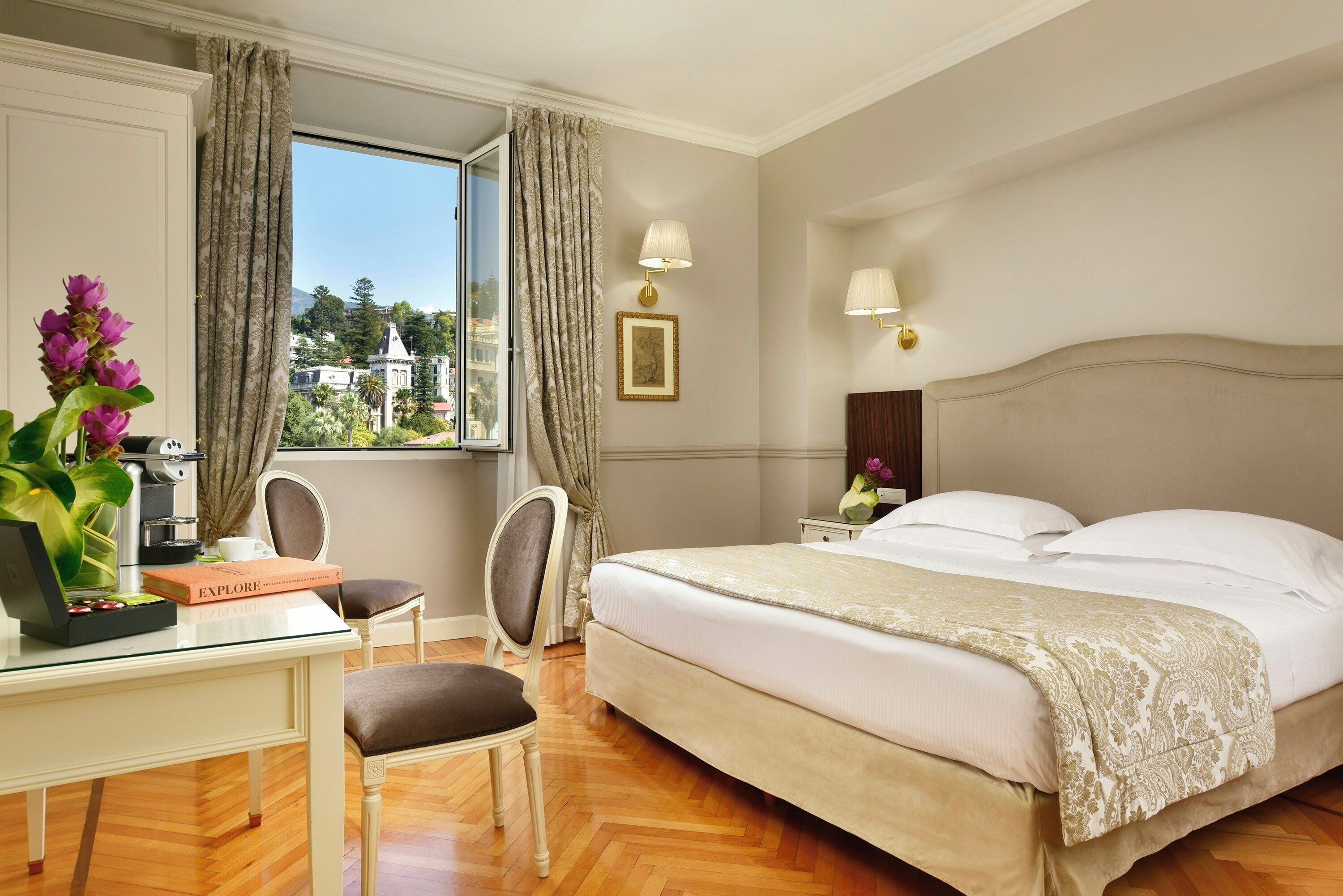 Une chambre d’hôtel avec un lit, un bureau, une chaise, une fenêtre avec des rideaux, une lampe et une vue sur l’extérieur.