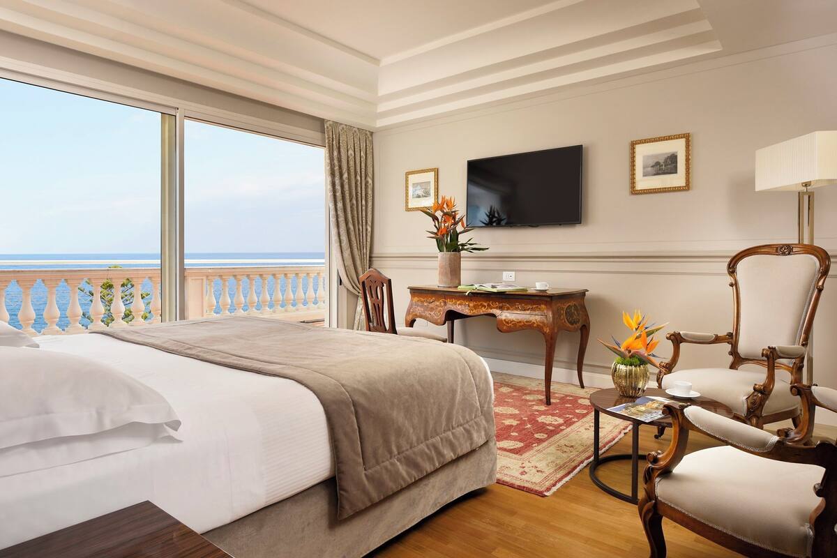 A hotel room with a large bed, a desk with a chair, a TV, and a view of the sea.