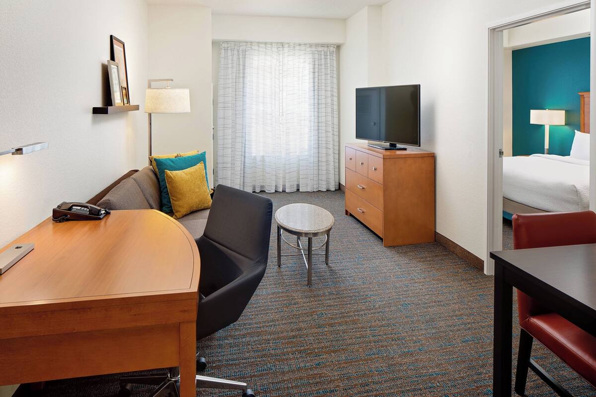 Habitación de hotel con sofá, una mesita redonda, televisión sobre un mueble de madera, cama con cabecera azul y ventana con cortinas.