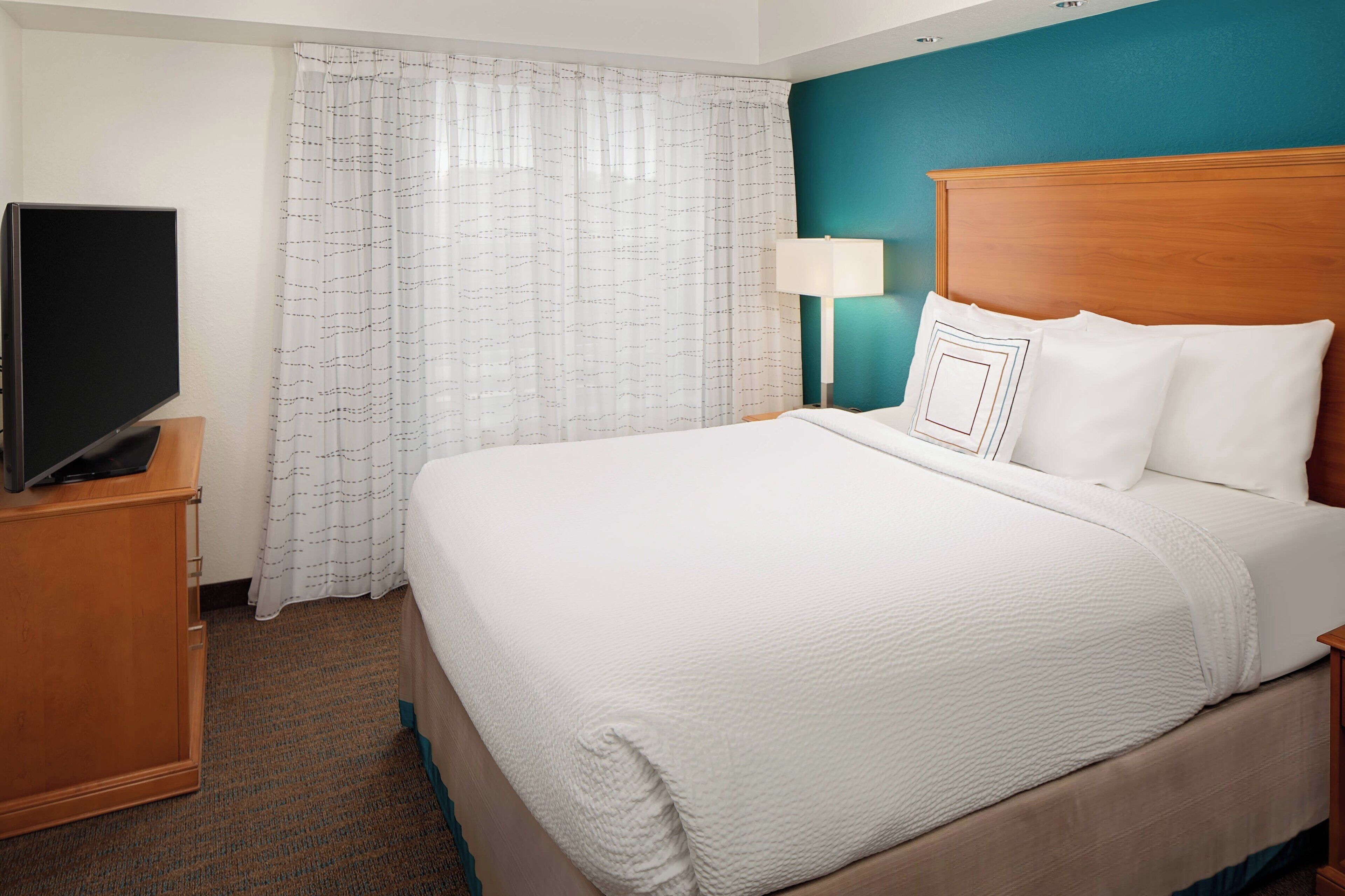 A hotel room with a bed, a TV on a wooden cabinet, a lamp, and a window with curtains.