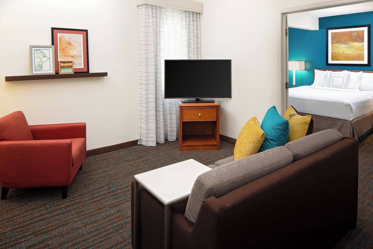 Habitación de hotel con sofá marrón, sillón rojo, mesa blanca, televisor de pantalla plana sobre un soporte de madera, cama con ropa blanca y estante empotrado con obras de arte enmarcadas.