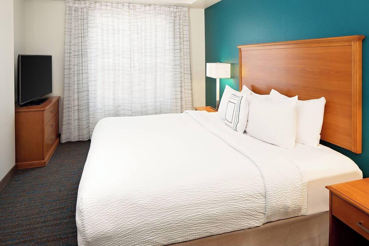 Habitación de hotel con una cama grande, un televisor sobre un mueble de madera, una mesita de noche con una lámpara y una ventana con cortinas.