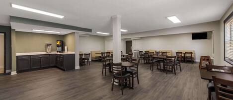 A spacious dining area with multiple tables and chairs, a kitchen area with dark cabinets, and a flat-screen TV mounted on the wall.