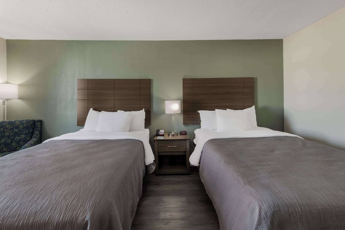 A hotel room with two beds, a nightstand, a lamp, and a chair.