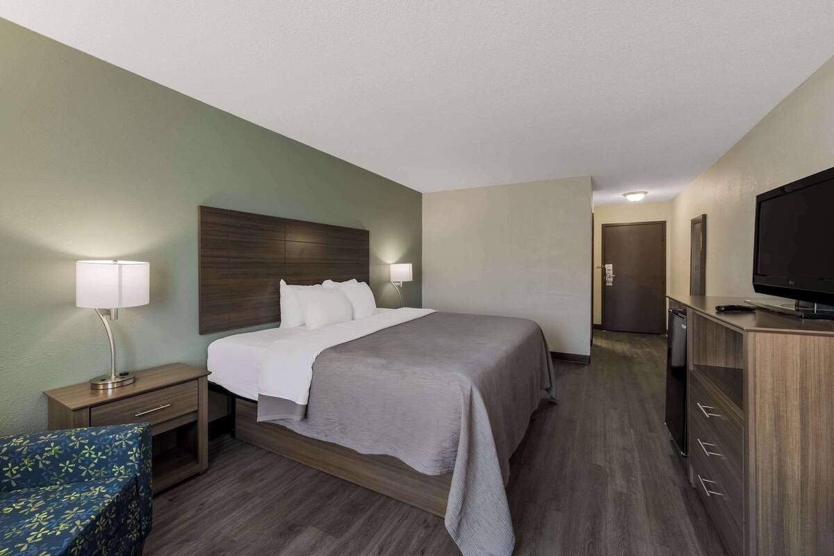 A hotel room with a bed, bedside table, lamp, TV, and a small seating area.