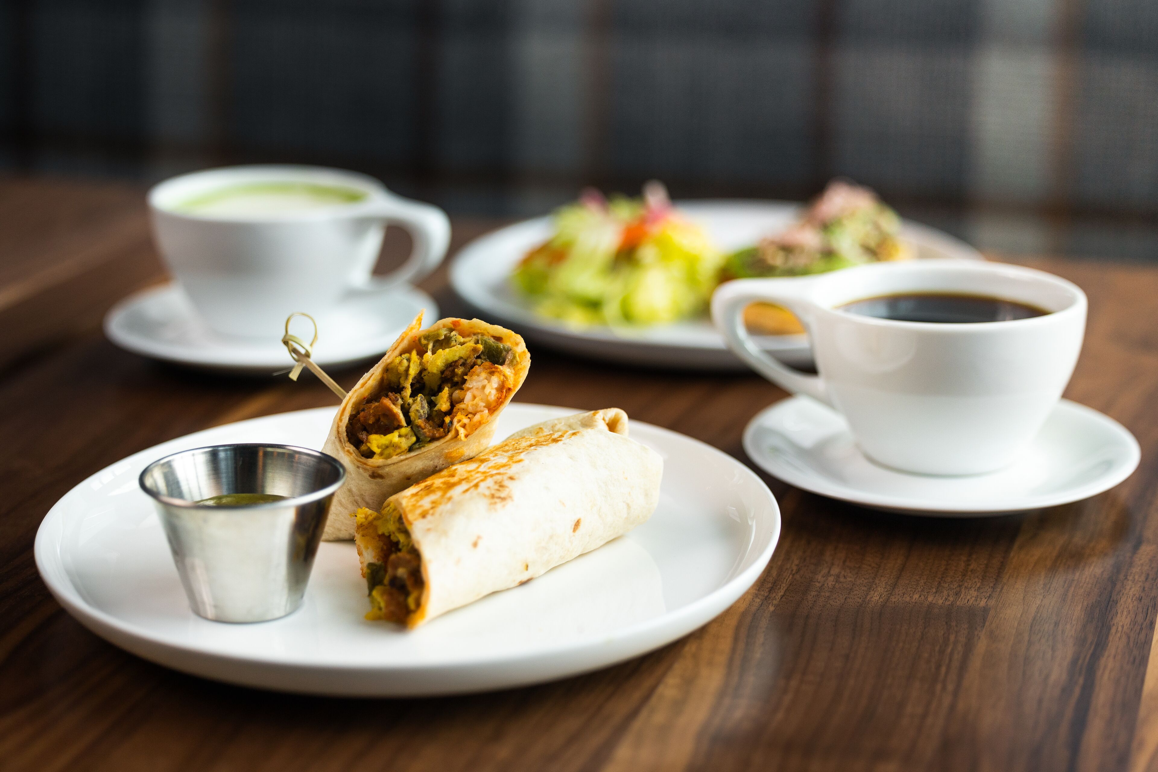 Una mesa con un burrito, una taza de café y un plato de ensalada.