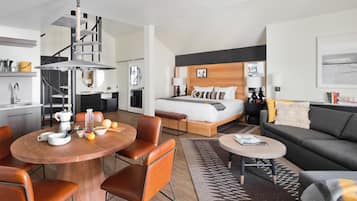 A modern hotel room with a bed, a dining area with a round table and chairs, a sofa, and a staircase.