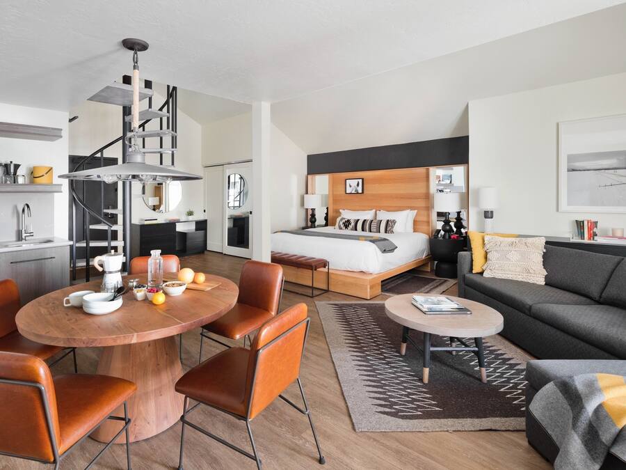 A modern hotel room with a bed, a dining area with a round table and chairs, a sofa, and a staircase.