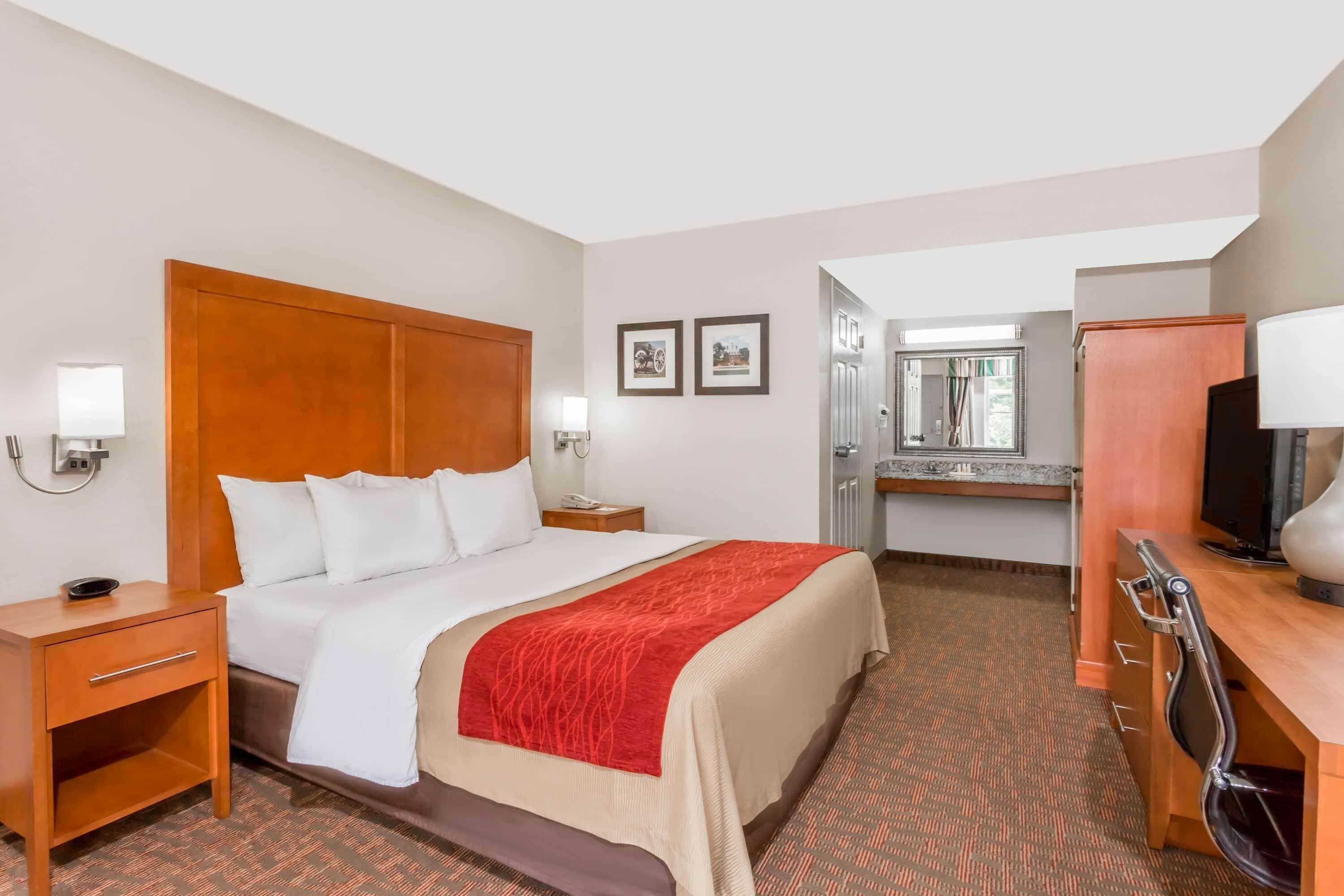 A hotel room with a large bed, a wooden nightstand, a desk with a chair, a television, and a mirror.