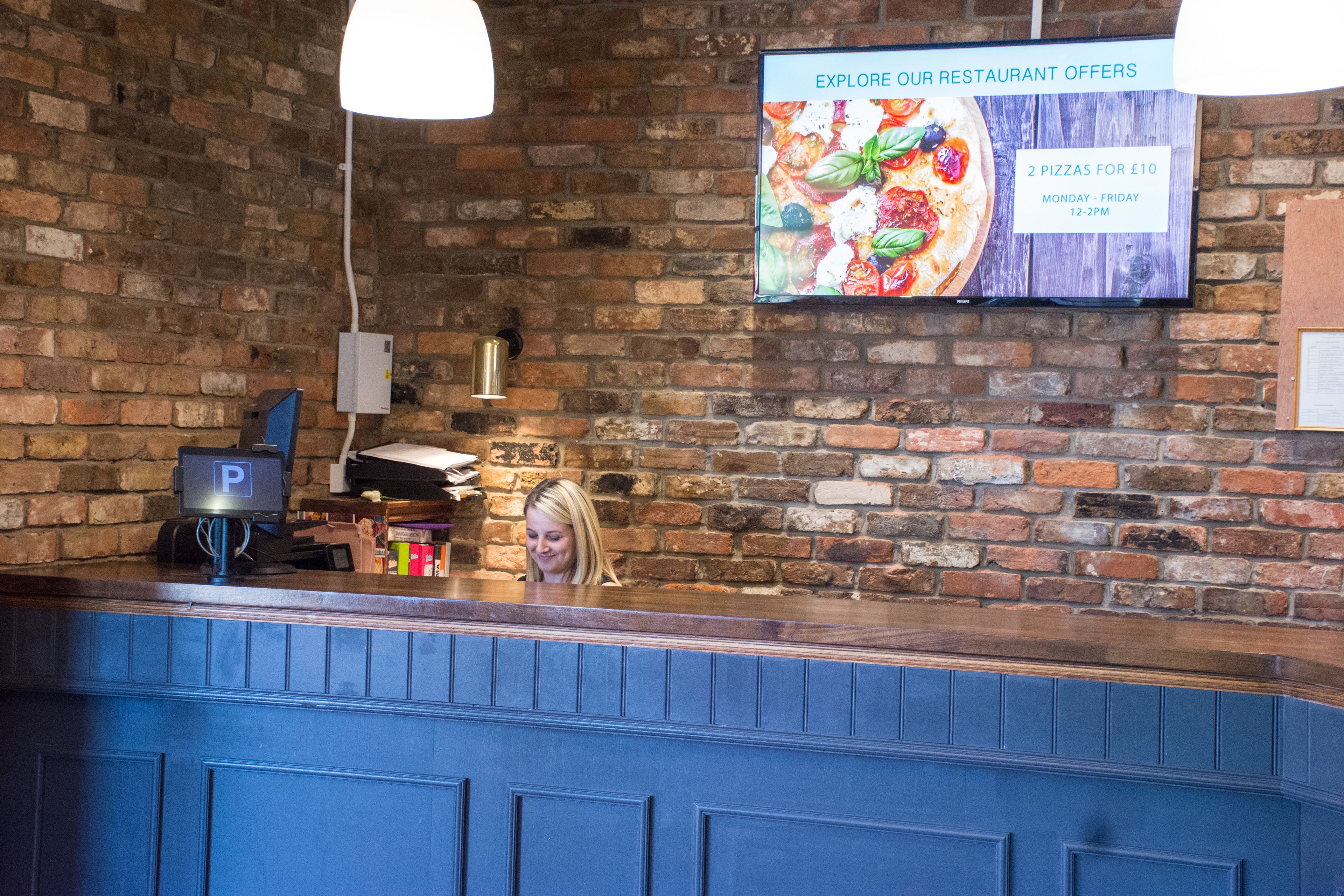 Un comptoir de restaurant avec un mur en briques en arrière-plan, un tableau affichant les offres du menu et un écran numérique présentant les plats proposés.