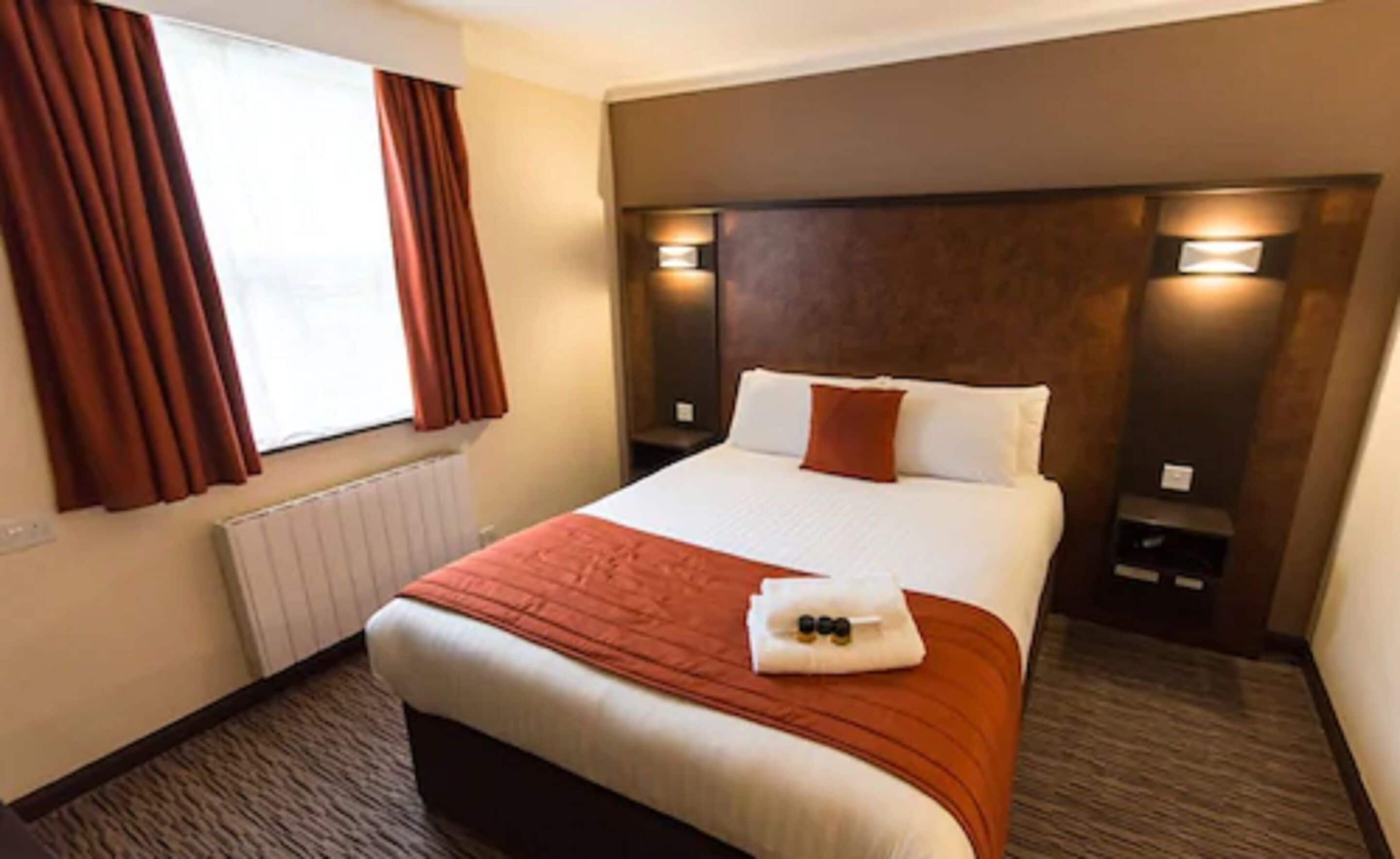 A hotel room with a large bed, red curtains, a radiator, and two wall-mounted lights.