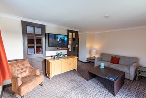 Habitación de hotel con una gran televisión de pantalla plana montada en la pared, un armario de madera, un sofá color beige, una mesa de centro y una mesita de noche con lámpara.