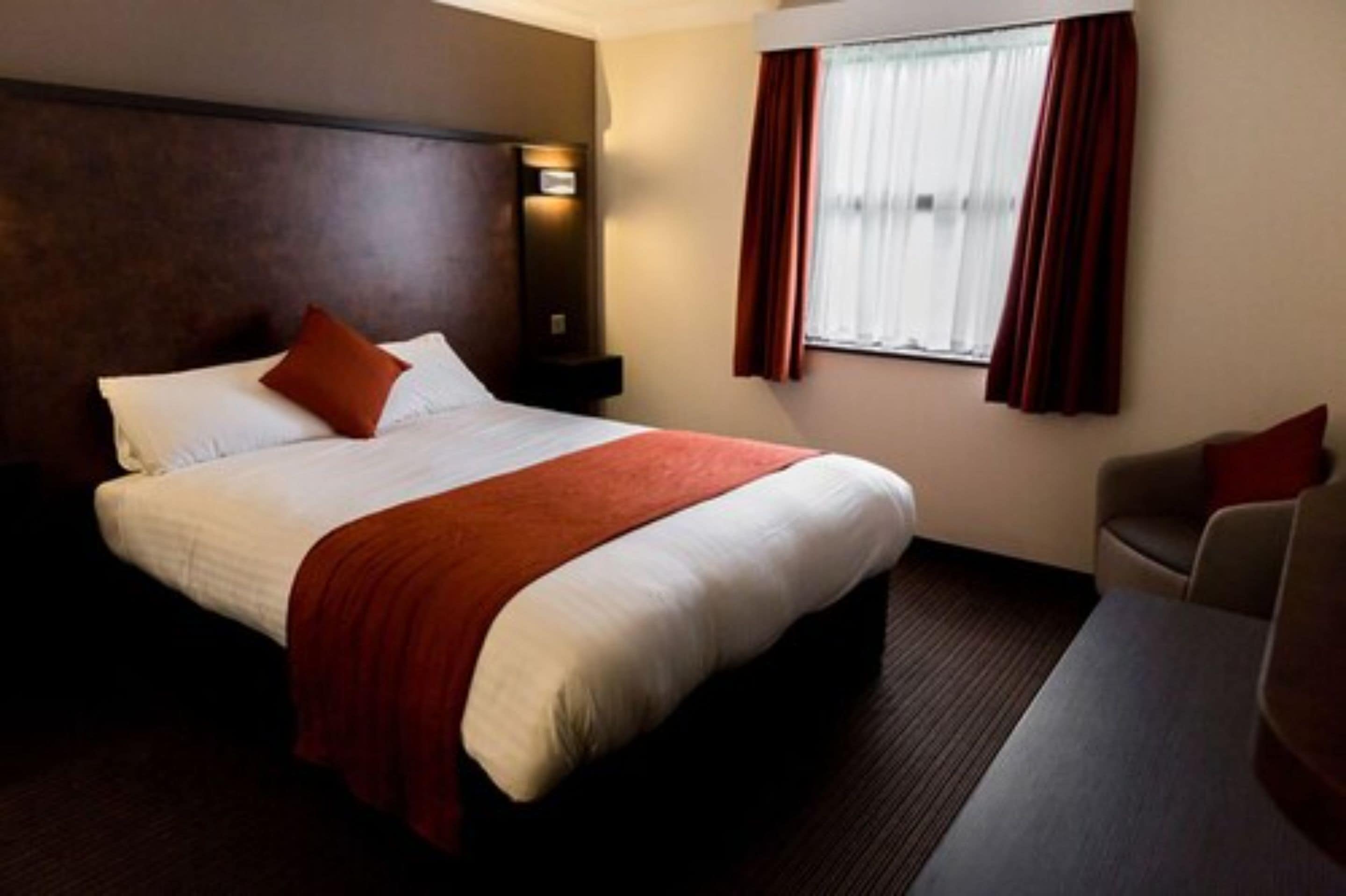 A hotel room with a large bed, a nightstand, a desk, and a window with curtains.
