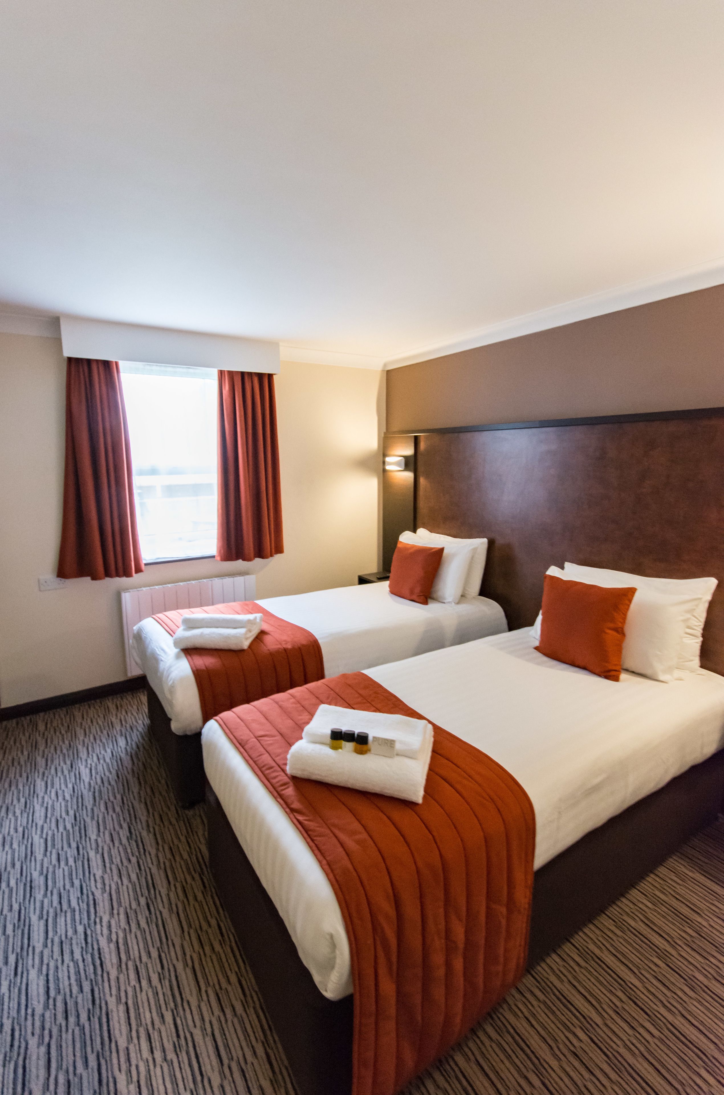 Une chambre d’hôtel avec deux lits, chacun agrémenté de linge de lit blanc et de touches de couleur orange. On y trouve une fenêtre avec des rideaux, une tête de lit et un sol recouvert de moquette.