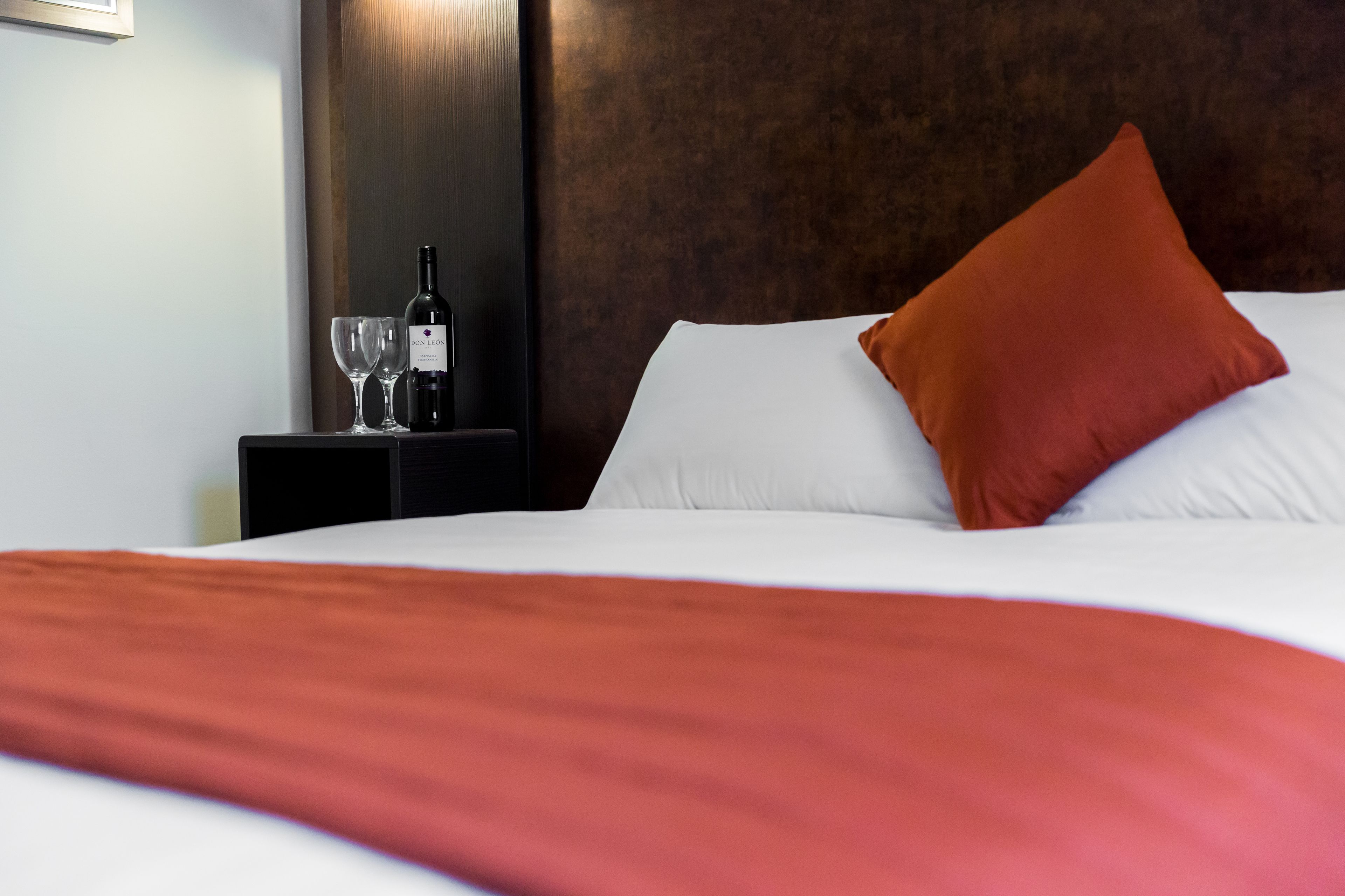 A neatly made bed with a white comforter and a red throw blanket, a brown pillow, and a bottle of wine on a bedside table with two glasses.