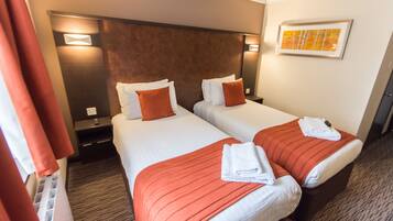 A hotel room with two beds, each with a red bedspread and white pillows, a nightstand with a lamp, and a framed picture on the wall.