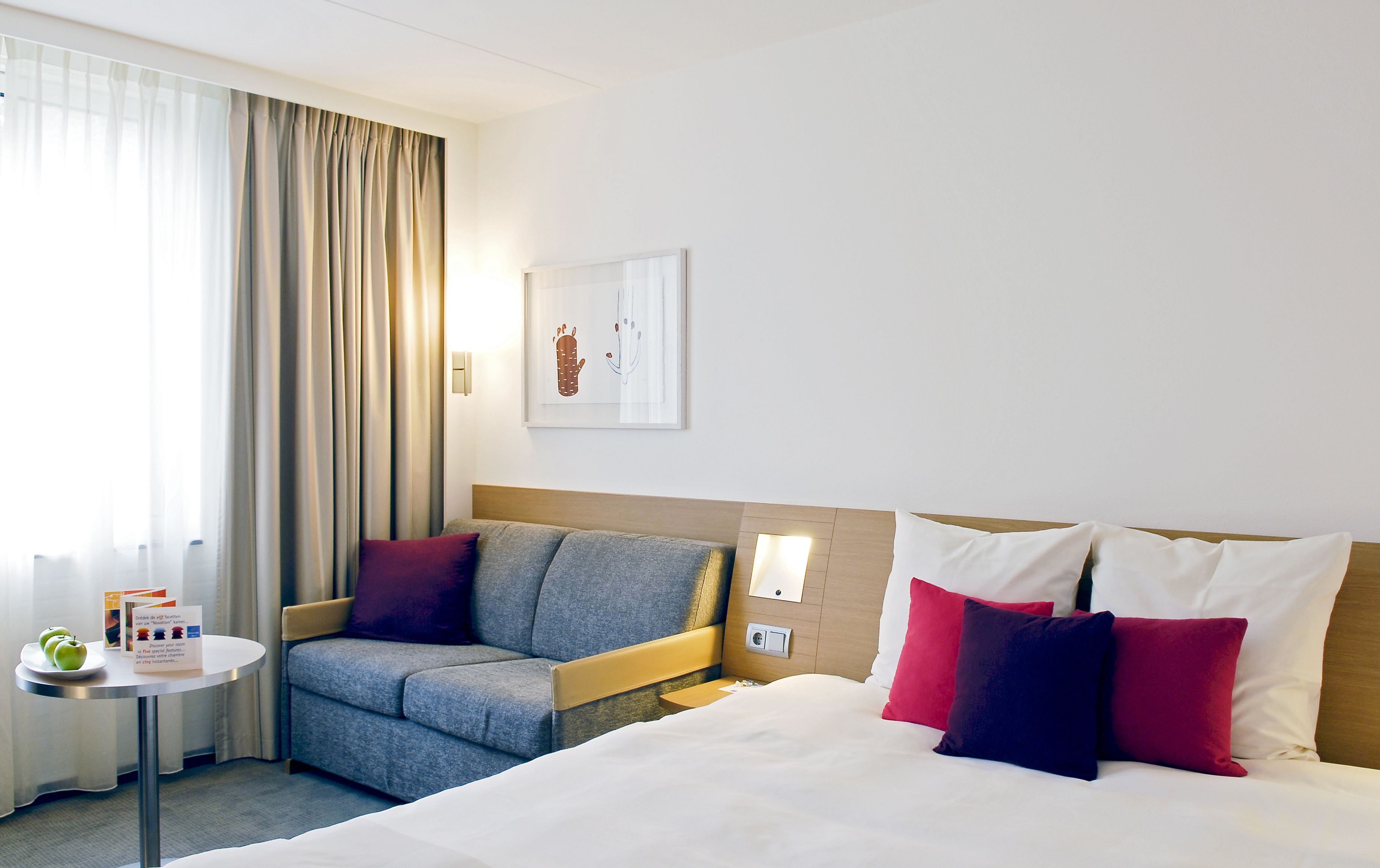 Ett hotellrum med en säng, en soffa, ett litet bord med en tallrik full av äpplen och ett fönster med gardiner.