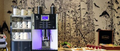 Una cafetera con varias opciones de preparación y una pantalla, frente a una pared con diseño de ramas de árbol.