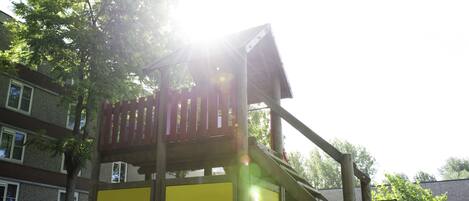 A wooden play structure with a slide and a platform, surrounded by trees and a building in the background.
