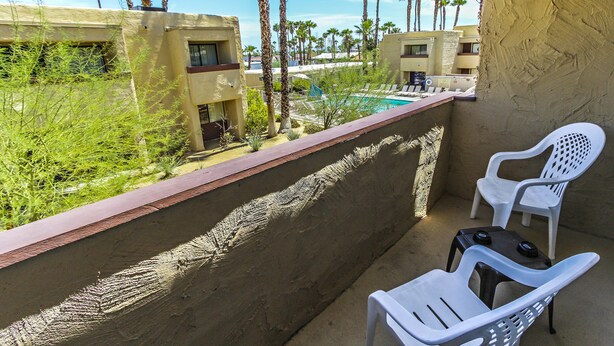 Ein Balkon mit zwei weißen Plastikstühlen, einem kleinen Tisch und Blick auf einen Poolbereich mit Palmen und Gebäuden im Hintergrund.
