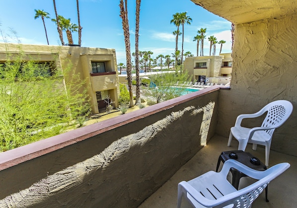 A balcony with two white plastic chairs, a small table, and a view of a pool area with palm trees and buildings in the background.