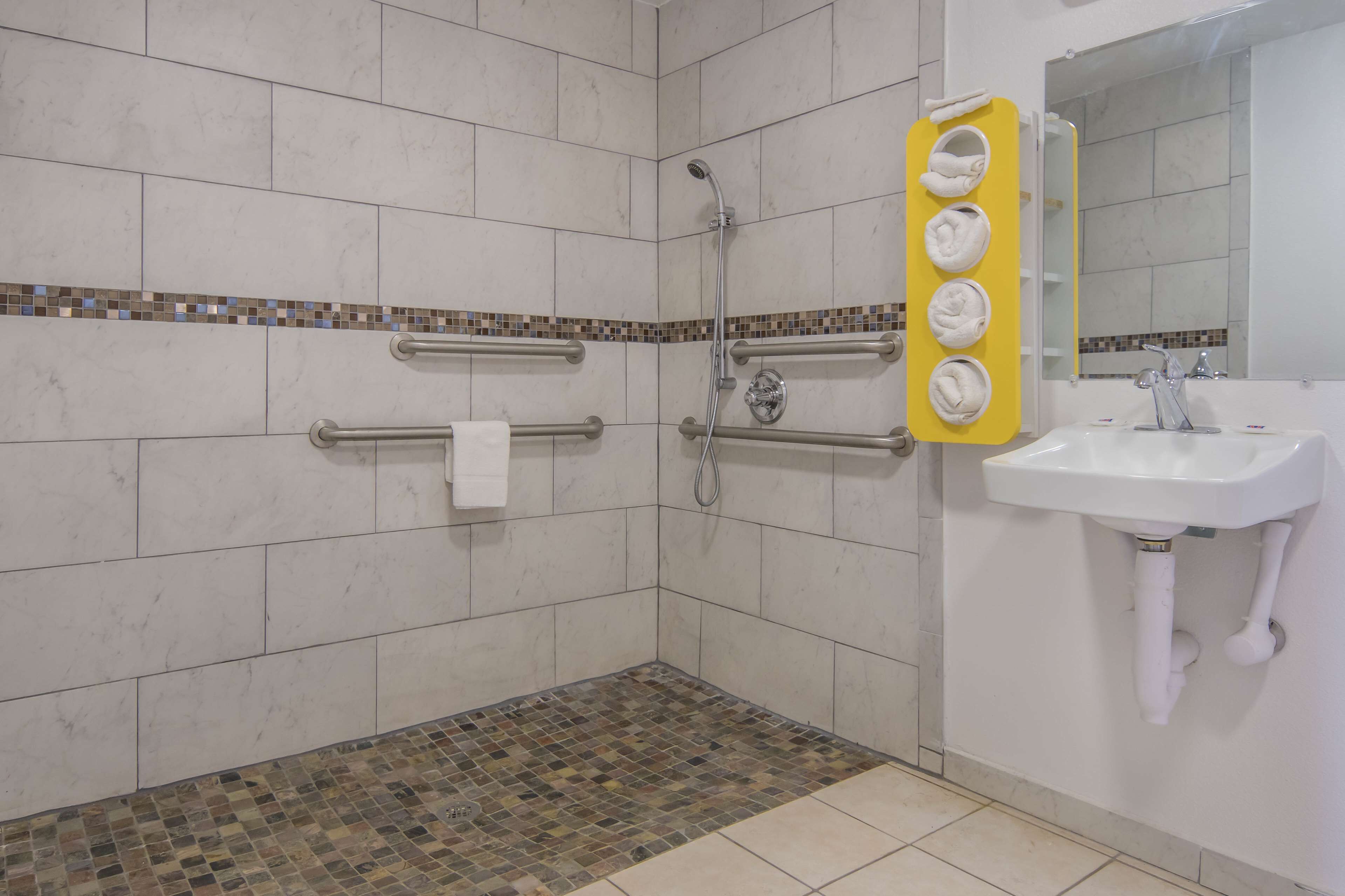 Een badkamer met witte tegels, een douche met handdouche, een wastafel met spiegel en een handdoekrek met handdoeken.