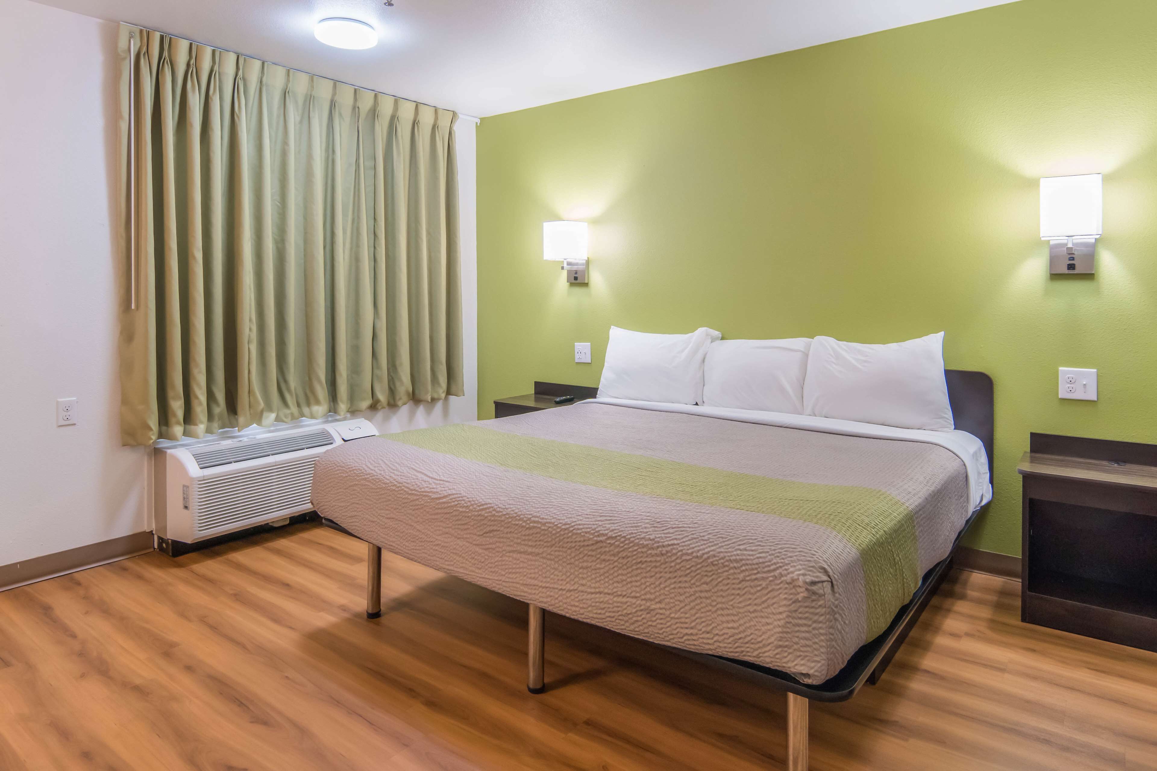 A hotel room with a bed, two bedside tables, a wall-mounted air conditioner, a window with curtains, and two wall-mounted lights.