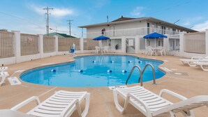 Una piscina con sillones de descanso y sombrillas, rodeada por una valla blanca y un edificio al fondo.