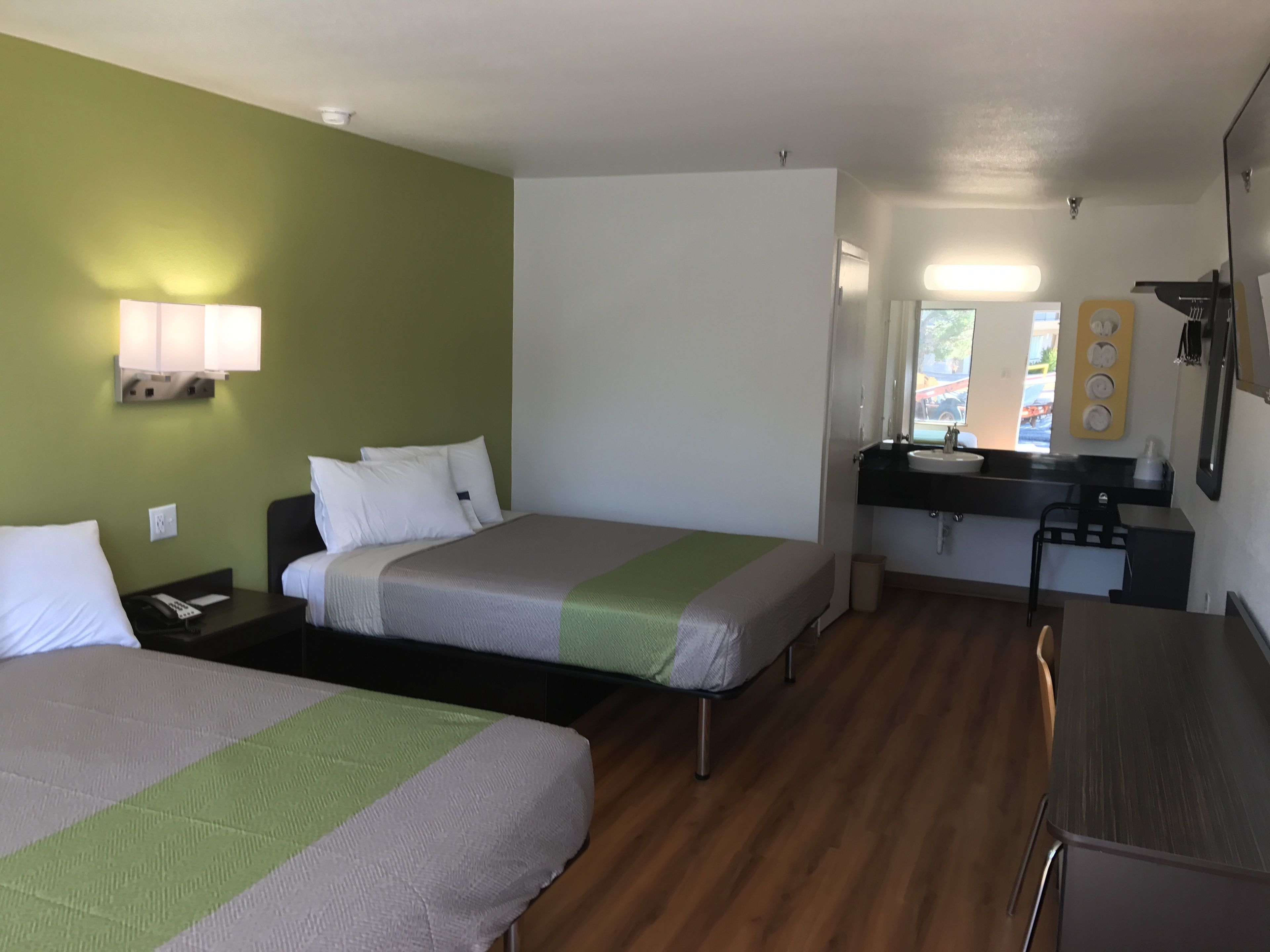 A hotel room with two beds, a small kitchenette, a mirror, and a window with a view of the outside.