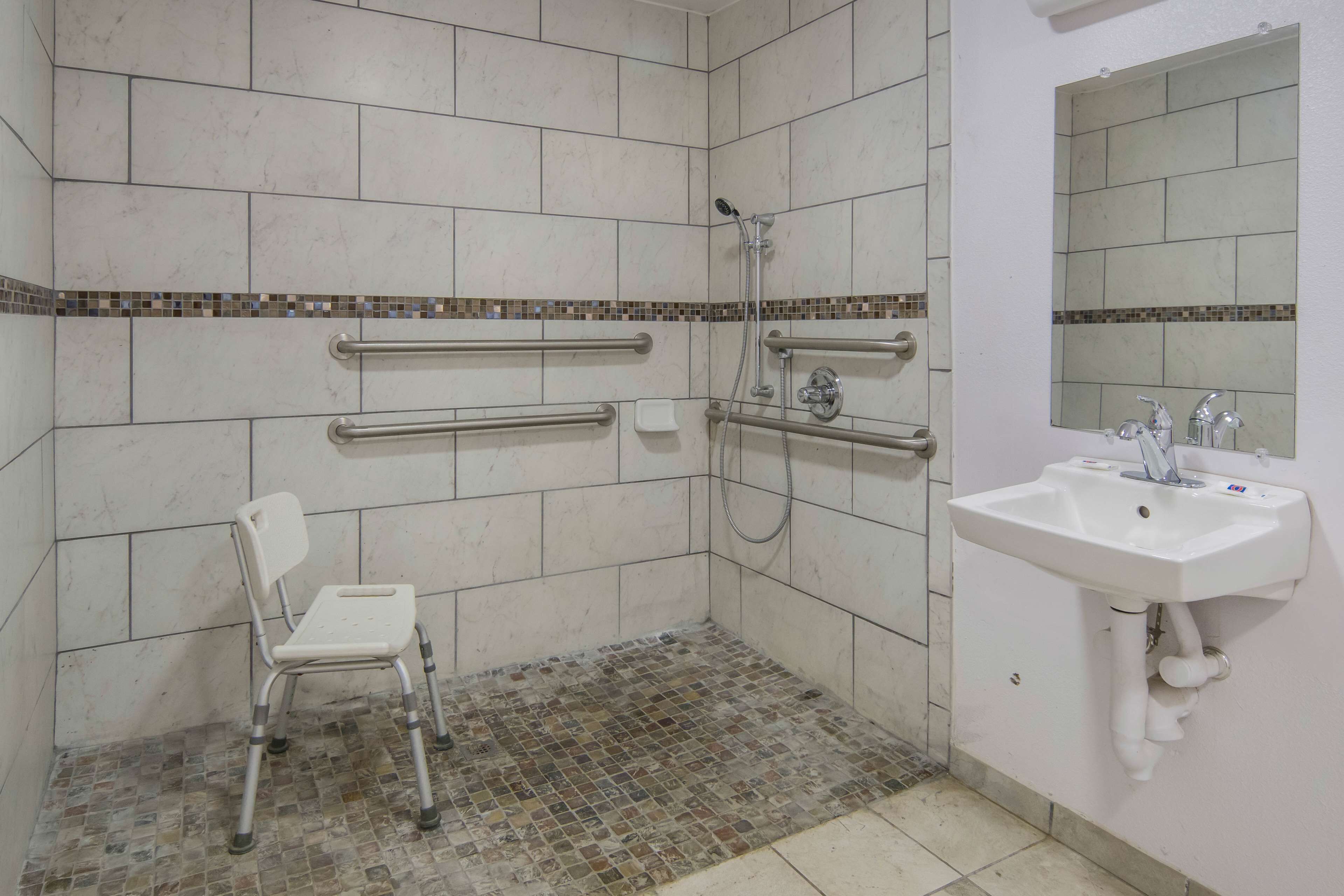 A bathroom with white tiles, a shower with a handheld showerhead, a white sink with a mirror, and a wheelchair-accessible bench.